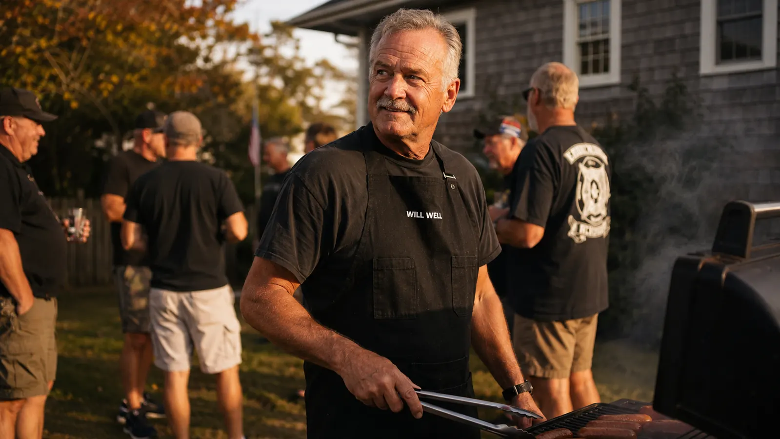 Retired firefighter in a black apron standing at a smoking grill in his backyard while a group of older men gather around him on a cool October afternoon