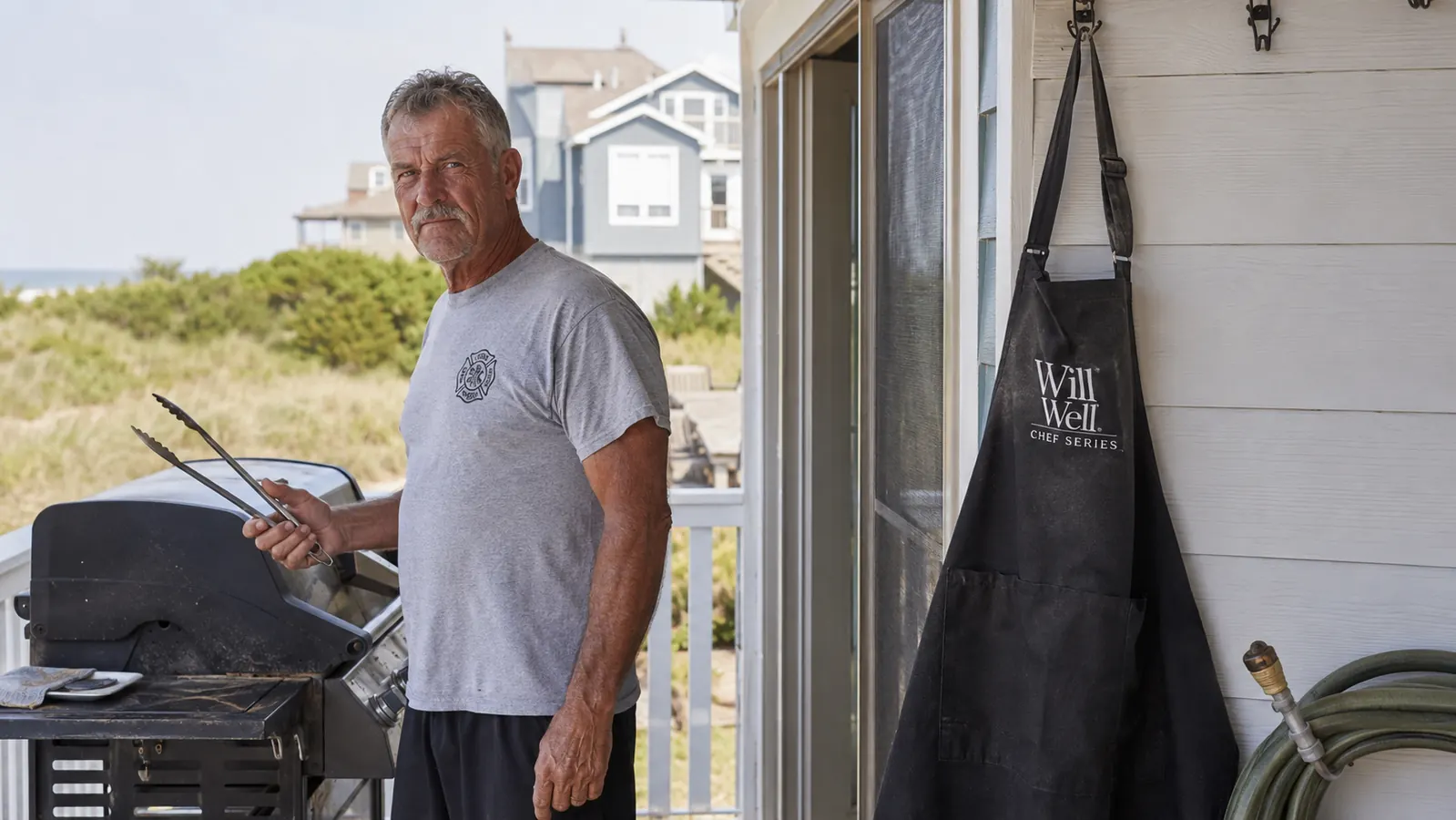 A worn black Will Well apron hanging on an exterior hook near a grill, sun-faded slightly across the shoulder strap